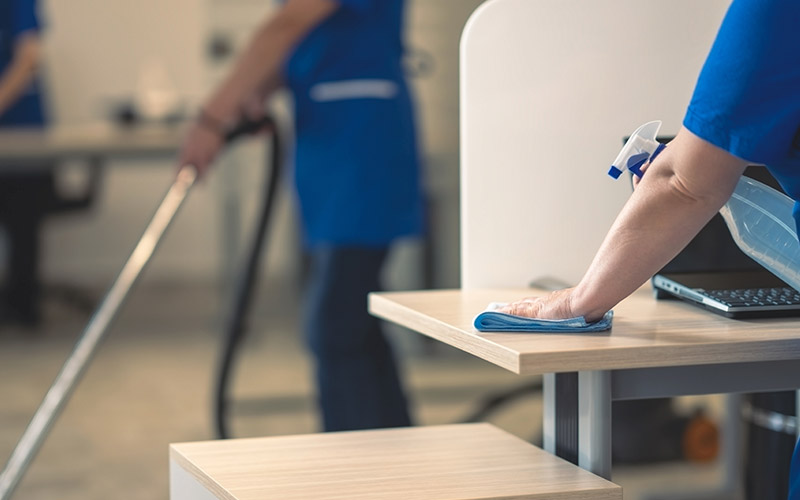 Professionelle Büroreinigung in Offenbach: Reinigungskraft säubert Schreibtischoberflächen und Arbeitsbereiche, während im Hintergrund der Boden gereinigt wird – hygienische Reinigung für Unternehmen und Büros.